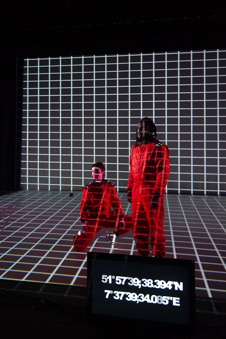 Zwei PerformerInnen in roten Overalls auf einer dunklen Bühne mit Rastermotiv