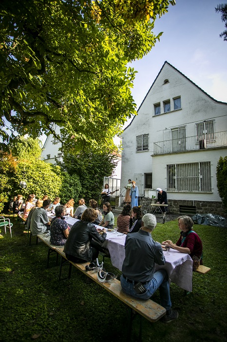 Eine Gruppe Theaterbesucher*innen sitzt im Garten eines Hauses an einr langen Tafel